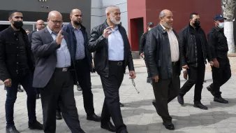 Nell'immagine si vede un gruppo di uomini che camminano insieme all'aperto. Alcuni di loro indossano giacche di pelle e altri abiti formali. Alcuni uomini indossano mascherine, mentre altri no. Sembrano impegnati in una conversazione e uno di loro alza la mano in un gesto di saluto. Sullo sfondo si intravedono strutture architettoniche moderne e una pavimentazione in pietra. L'atmosfera sembra formale e organizzata.