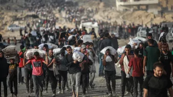 Gruppo di persone che trasportano sacchi su un terreno polveroso, in un contesto affollato.