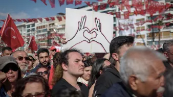 L'immagine mostra una grande folla riunita in un evento all'aperto, probabilmente una manifestazione o una celebrazione. Alcuni partecipanti sostengono bandiere turche, che sono di un rosso acceso, mentre un uomo in primo piano è visibile con una maglietta nera e guarda verso la telecamera. Dietro di lui, c'è un cartello che rappresenta due mani che formano un cuore, simbolo di amore o unità. L'atmosfera sembra essere festosa e piena di energia, con molte persone che condividono un momento di comunità.