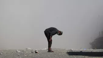 Un uomo anziano si piega in avanti su una strada polverosa, circondato da detriti.