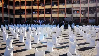 L'immagine mostra un'area all'aperto con molti sacchi bianchi disposti in file ordinate. Sullo sfondo si vede un edificio di più piani, con diversi balconi. Alcuni panni sono appesi ad asciugare tra i balconi. La scena è illuminata dalla luce del sole, creando un'atmosfera vivace e attiva. Il pavimento è costituito da mattonelle in cui si possono notare i segni del tempo. L'insieme suggerisce un contesto urbano con attività legate alla distribuzione di beni.