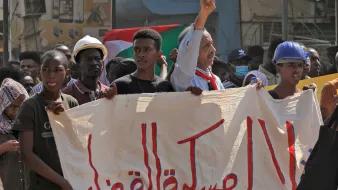 L'immagine mostra una manifestazione con diverse persone che sollevano cartelli e bandiere. In primo piano, un uomo con un cappello e una sciarpa al collo alza la mano mostrando il simbolo della pace. Gli altri manifestanti sembrano esprimere supporto per una causa, cominciando da una grande bandiera sudanese. Lo sfondo mostra dei cartelloni pubblicitari, mentre l'atmosfera appare carica di energia e determinazione.