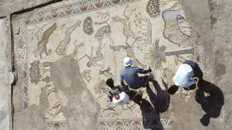 L'immagine mostra un antico mosaico, parzialmente scavato, con una varietà di animali e motivi decorativi. Si possono osservare figure di leoni, cervi e palme, che suggeriscono una rappresentazione di scene naturali. Due persone sono visibili nella parte inferiore, impegnate nel lavoro di pulizia o restauro del mosaico. L'illuminazione crea ombre sul pavimento, accentuando i dettagli del mosaico e l'atmosfera archeologica del sito.