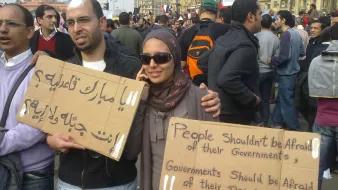 L'immagine mostra un gruppo di persone in una manifestazione. Due individui in primo piano tengono in mano dei cartelli. Uno dei cartelli è scritto in arabo e l'altro in inglese. Il messaggio in inglese dice "Le persone non dovrebbero avere paura dei loro governi. I governi dovrebbero avere paura del loro popolo." I partecipanti sembrano impegnati e determinati, con un'ampia folla di persone sullo sfondo. La scena evoca un senso di protesta e richiesta di cambiamento sociale.