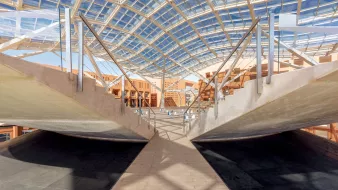 L'immagine mostra un'architettura moderna caratterizzata da una struttura ampia e luminosa. La copertura in vetro permette alla luce naturale di filtrare, creando un ambiente aperto e arioso. Le pareti laterali presentano un design minimalista, con elementi di legno e cemento che si combinano armoniosamente. Al centro, una passerella in legno collega le diverse aree, accentuando una sensazione di continuità e apertura nello spazio. La scena è caratterizzata da linee pulite e forme geometriche, suggerendo un design innovativo e funzionale.