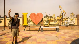 Un uomo fa un selfie davanti a un grande cartello colorato con "I ❤️" e fiori.