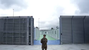 L'immagine mostra un soldato in uniforme, in piedi al centro di un'area delimitata. Sullo sfondo si possono vedere due strutture metalliche grigie che sembrano essere recinzioni, con un'area di pavimentazione colorata di verde e blu tra di esse. Il cielo è nuvoloso, suggerendo una giornata grigia. L'atmosfera sembra essere di sicurezza e controllo, con un forte senso di vigilanza da parte del soldato.