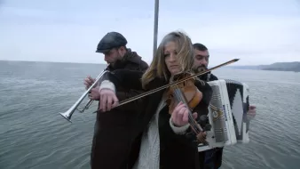 L'immagine mostra tre musicisti che suonano su una piattaforma sopra l'acqua. Una giovane donna al centro suona il violino, mentre indossa un maglione chiaro e un cappotto scuro. A sinistra c'è un uomo che suona un flauto ed è vestito con un cappello e un cappotto scuro. A destra, un altro uomo suona l'accordeon. Lo sfondo è caratterizzato da acque calme e un cielo nuvoloso, creando un'atmosfera suggestiva e malinconica.