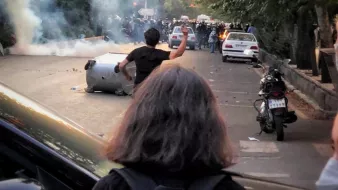 L'immagine mostra una scena di tensione e conflitto in una strada. Ci sono persone in piedi, alcune sembrano protestare, e si nota un'atmosfera di caos con fumi o gas lacrimogeni nell'aria. Alcuni veicoli sono coinvolti, con una macchina ribaltata, mentre altri autoveicoli sono parcheggiati lungo il marciapiede. Gli alberi ai lati della strada contribuiscono a dare un'idea di una zona urbana. La persona in primo piano, che ha i capelli lunghi, sembra stia alzando un braccio verso qualcosa, forse in segno di protesta o sfida.