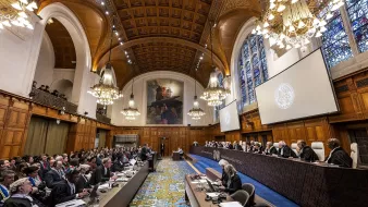L'immagine mostra un'aula di giustizia, probabilmente una corte internazionale. La sala è caratterizzata da un'architettura imponente con soffitti alti, lampadari eleganti e finestre ad arco che lasciano filtrare la luce. In primo piano, c'è un lungo tavolo con diversi avvocati e rappresentanti, mentre sul lato opposto, una serie di giudici sono seduti su una piattaforma rialzata. Il pubblico è composto da molte persone, alcune delle quali prendono appunti. L'atmosfera sembra formale e rispettosa, tipica di un'importante udienza legale.