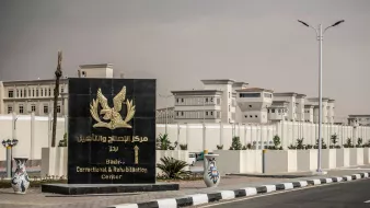 L'immagine mostra un grande cartello di benvenuto che indica il "Centro di Correzione e Riabilitazione di Badia". Il cartello è di colore scuro con un simbolo o un emblema, e si trova all'ingresso di una struttura recintata. Sullo sfondo si possono vedere edifici moderni, circondati da piante e un'illuminazione stradale. Il contesto appare ordinato e ben tenuto, suggerendo un'area istituzionale.