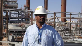 L'immagine mostra un uomo che indossa un casco di sicurezza bianco e una camicia azzurra. Si trova in un ambiente industriale, con impianti e tubazioni sullo sfondo. L'atmosfera sembra quella di un sito di produzione o di una raffineria, con diverse strutture metalliche e torri visibili. L'uomo appare fiducioso e professionale mentre si pone di fronte all'obiettivo.