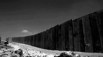 L'immagine mostra un muro lungo e imponente, eretto in un paesaggio desolato. La scena è in bianco e nero, il che le conferisce un'atmosfera drammatica. Il muro, costituito da grandi pannelli di cemento, si estende in modo curvilineo verso l'orizzonte, mentre il terreno attorno appare roccioso e poco curato. In lontananza, si intravede un cielo chiaro, con una nube che si staglia sullo sfondo, aggiungendo profondità all'immagine. L'assenza di colori enfatizza la severità e la rigidità della struttura, suggerendo un senso di divisione e separazione.