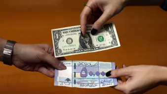 L'immagine mostra due mani che si scambiano delle banconote. Una mano tiene una banconota da un dollaro statunitense, mentre l'altra mano presenta una banconota di una valuta diversa, probabilmente una banconota di un paese arabo. Il fondo sembra essere un tavolo di legno, creando un contrasto con le banconote in primo piano.