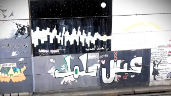 L'immagine mostra un murale colorato su un muro, con diversi disegni e scritte in arabo. In primo piano, si legge "عيش حلمك" che significa "Vivi il tuo sogno". Lo sfondo presenta vari elementi artistici, tra cui silhouette di grattacieli e figure stilizzate, creando un'atmosfera urbana e vibrante. Ci sono anche elementi decorativi e simboli che riflettono un messaggio di speranza e ispirazione.
