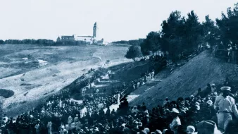 Un grande raduno di persone all'aperto, con un edifico distante e alberi circostanti.