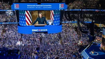 L'immagine mostra una scena di una grande convention politica, probabilmente la Convenzione Nazionale Democratica del 2024 a Chicago. La sala è affollata di persone che applaudono, con molti sostenitori che sventolano bandiere e cartelli. Al centro, un grande schermo mostra un discorso di una figura politica (probabilmente un leader del partito). Gli ambienti sono decorati con colori e simboli del partito, come stelle e strisce. L'atmosfera sembra essere di entusiasmo e impegno politico.