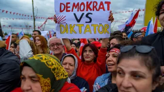 Nell'immagine si vede un gruppo di persone ad una manifestazione. In primo piano, una donna sorridente indossa una maglietta rossa e tiene in mano un cartello con la scritta "DEMOCRAT VS DIKTATÖR". Intorno a lei, ci sono diverse altre persone che sembrano partecipare attivamente all'evento. Molti di loro indossano sciarpe e cappelli colorati. Sullo sfondo, si vedono bandiere e decorazioni che contribuiscono all'atmosfera di protesta. L'ambientazione sembra carica di emozioni e impegno civico.