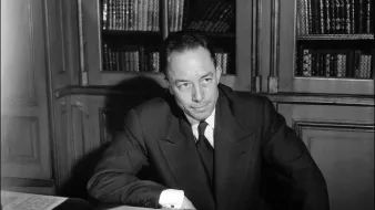 L'immagine mostra un uomo seduto a un tavolo, con dei documenti sparsi davanti a lui. Indossa un abito scuro con una cravatta, e appare pensieroso mentre guarda verso un punto oltre la fotocamera. Sullo sfondo si intravedono alcune librerie con libri. L'illuminazione è elegante, creando un'atmosfera seria e riflessiva.