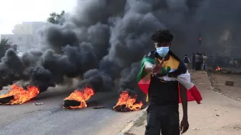 L'immagine mostra un giovane che cammina lungo una strada, con fumi neri che si alzano dietro di lui. Ci sono pneumatici in fiamme che bruciano ai lati della strada. Il ragazzo indossa una mascherina e fa un gesto di pace con la mano. Sullo sfondo, si notano altre persone che osservano la scena. L'atmosfera sembra intensa e carica di tensione.