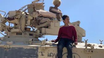 L'immagine mostra un giovane ragazzo seduto su un veicolo militare, apparentemente un carro armato. Accanto a lui c'è una figura che sembra essere una bambola o un manichino con capelli biondi. Sul carro armato è visibile una scritta che dice "I love you". Il cielo è limpido e blu, creando un contrasto con l'ambiente circostante. Il ragazzo indossa un maglione rosso e jeans, mentre la bambola è posizionata in modo da sembrare tranquilla e serena.