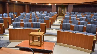 L'immagine mostra un'aula legislativa vuota, caratterizzata da una disposizione ordinata di banchi in legno con sedute nere. Al centro della scena si trova una piccola struttura in legno, probabilmente un tavolo o una cassa. Le pareti dell'aula sono di un colore scuro, mentre il pavimento è di tonalità più chiara, creando un contrasto. L'ambiente sembra formale e istituzionale, suggerendo un luogo di discussione e decisione politica.