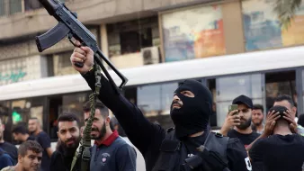 L'immagine mostra un uomo mascherato che brandisce un fucile automatico in una strada affollata. Si possono vedere altre persone intorno a lui, alcune delle quali sembrano sostenere o osservare l'azione. L'ambiente circostante è urbanizzato, con edifici e un autobus che appare in lontananza. L'atmosfera sembra carica di tensione, suggerendo un contesto di protesta o conflitto.