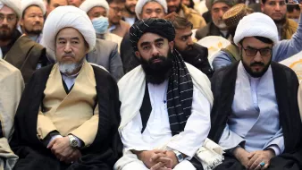 L'immagine mostra un gruppo di uomini seduti in un contesto di evento o incontro. I partecipanti indossano abiti tradizionali, tra cui chador e turbanti. I volti esprimono varie emozioni, alcuni sembrano seri e pensierosi. Sullo sfondo si possono vedere altre persone, suggerendo che si tratta di un'assemblea o di una manifestazione. L'atmosfera sembra essere formale e rispettosa.