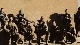 L'immagine mostra un gruppo di otto soldati in divisa militare, affiancati e in posa. Sono armati e sembrano essere in un ambiente desertico o in una zona di conflitto. I soldati indossano attrezzature tattiche e occhiali da sole, e la luce evidenzia i dettagli delle loro uniformi. Sullo sfondo si vede una parete di un edificio, suggerendo un contesto operativo. L'atmosfera è seria e professionale.