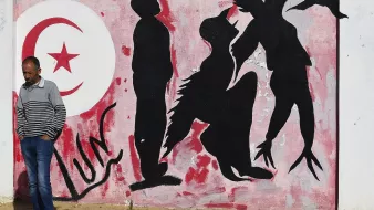 L'immagine mostra un murale su un muro bianco, caratterizzato da una palette di colori rossi e neri. Sullo sfondo, è presente un grande simbolo della luna crescente e una stella, simboli spesso associati alla bandiera tunisina. Al centro, ci sono delle silhouette di figure umane che appaiono in diverse posizioni: alcune sono in piedi, altre sembrano inginocchiarsi o muoversi in modo dinamico. In primo piano, un uomo con una maglietta a strisce passeggia accanto al murale, creando un contrasto tra l'arte e la vita quotidiana. L'atmosfera trasmette un senso di cultura e identità, evidentemente legato alla Tunisia.