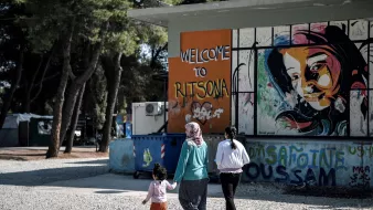 L'immagine mostra tre donne che stanno camminando in una zona all'aperto. Una di loro tiene per mano una bambina, mentre un'altra donna indossa un hijab. Sullo sfondo si può vedere un murale colorato che rappresenta un volto di donna, accompagnato dalla scritta "Welcome to Ritsona". L'area sembra essere informale e accogliente, con alberi che la circondano e strutture varie visibili.