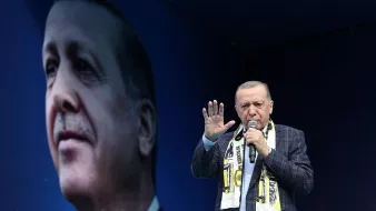 Nell'immagine si vede un uomo che parla a un pubblico, indossando una giacca a quadri e una sciarpa. Dietro di lui c'è un grande murale o un'immagine di un volto che lo ritrae. L'ambientazione sembra essere un evento pubblico o un comizio, con un'atmosfera di fervore e partecipazione. Il fondo è di colore blu, creando un contrasto con il soggetto principale.