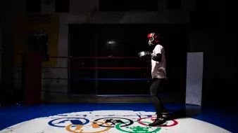 L'immagine mostra un giovane pugile che indossa i guantoni e un casco protettivo, mentre si allena in un ring da boxe. Lo sfondo è parzialmente illuminato, mettendo in risalto il pugile in azione. Sotto di lui, si trova un logo con i simboli olimpici e la scritta "ELBARRIO", che suggerisce un ambiente dedicato allo sport e alla formazione. La scena ha un'atmosfera di concentrazione e determinazione.