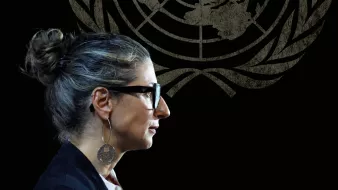 Donna con occhiali e capelli raccolti, in un ambiente con il logo delle Nazioni Unite sullo sfondo.
