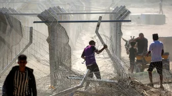 Uomini vicino a una recinzione di filo spinato in un'area polverosa e confusa.