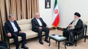 L'immagine mostra un incontro tra tre uomini. Due di loro sono seduti su un divano, mentre il terzo, che indossa un abbigliamento tradizionale, è seduto di fronte a loro. Sul muro si trova un ritratto e, accanto, è presente una bandiera iraniana. L'atmosfera sembra formale e il gruppo appare impegnato in una discussione.