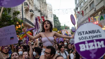 L'immagine mostra un gruppo di persone in una manifestazione per i diritti, con un forte focus sulle tematiche femministe e LGBTQ+. Una giovane donna, in primo piano e con i capelli ricci, è in piedi sulla spalle di un'altra persona e grida, esprimendo la sua passione. Molti partecipanti, prevalentemente donne, sono circondati da bandiere e cartelli colorati, alcuni con slogan che richiamano l'attenzione su temi di uguaglianza e diritti umani. L'atmosfera è vibrante e carica di energia, con un chiaro senso di solidarietà tra i manifestanti.