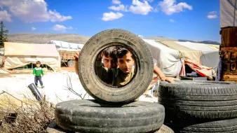 Nell'immagine si vedono due bambini che si affacciano attraverso un copertone di gomma, posto in orizzontale. I bambini, che sembrano curiosi e giocosi, sono circondati da altri copertoni impilati. Sullo sfondo si possono intravedere tende bianche e un paesaggio collinare sotto un cielo blu con alcune nuvole. L'atmosfera sembra essere di gioco e scoperta, nonostante l'ambiente circostante possa suggerire condizioni di vita difficili.