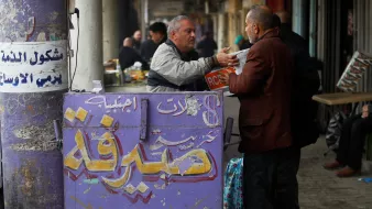 L'immagine mostra una scena di vita quotidiana in un mercato. Ci sono due uomini che interagiscono; uno sembra stia scambiando qualcosa con l'altro, mentre un'insegna colorata in arabo e inglese, che indica un'agenzia di cambio, è visibile in primo piano. Lo sfondo è caratterizzato da colonne e da un'atmosfera vivace tipica dei mercati affollati. Le persone e le attività circostanti suggeriscono un ambiente commerciale attivo.