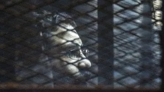 L'immagine mostra un uomo visto attraverso una rete metallica. La luce crea ombre sul suo viso, evidenziando la sua espressione seria. Indossa occhiali e ha una barba, mentre lo sfondo risulta scuro e indistinto, contribuendo a un'atmosfera di introspezione o solitudine.