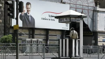 L'immagine mostra una cabina di polizia del traffico situata in una strada. In secondo piano, c'è un grande manifesto che ritrae un uomo che saluta, con il testo in arabo e italiano che dice "I Believe in Syria". La scena è animata ma appare tranquilla, con poche auto visibili e un'illuminazione chiara. Il contrasto tra la cabina di polizia e il manifesto può suggerire elementi di sicurezza e comunicazione politica.