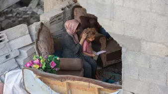 Due persone sedute su un divano in un ambiente distrutto, circondate da macerie.