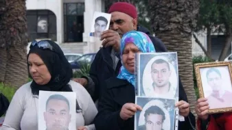Nell'immagine si vede un gruppo di persone che partecipano a una manifestazione. Alcuni di loro tengono in mano delle fotografie di uomini. Le donne indossano il velo e mostrano espressioni serie, mentre gli uomini dietro di loro sembrano sostenere le stesse fotografie. L'ambiente intorno è urbano, con alberi e edifici visibili sullo sfondo. La scena trasmette un senso di protesta e richiesta di giustizia.