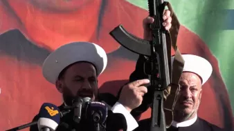 L'immagine mostra due uomini in abiti tradizionali, entrambi con copricapi bianchi, che sembrano partecipare a un evento pubblico o a una manifestazione. Uno di loro tiene in mano un fucile d'assalto, mentre l'altro osserva. Sullo sfondo c'è un grande murale o una bandiera che potrebbe rappresentare simboli politici o culturali. L'atmosfera sembra intensa e carica di emozione, con un focus sulla figura che tiene l'arma.