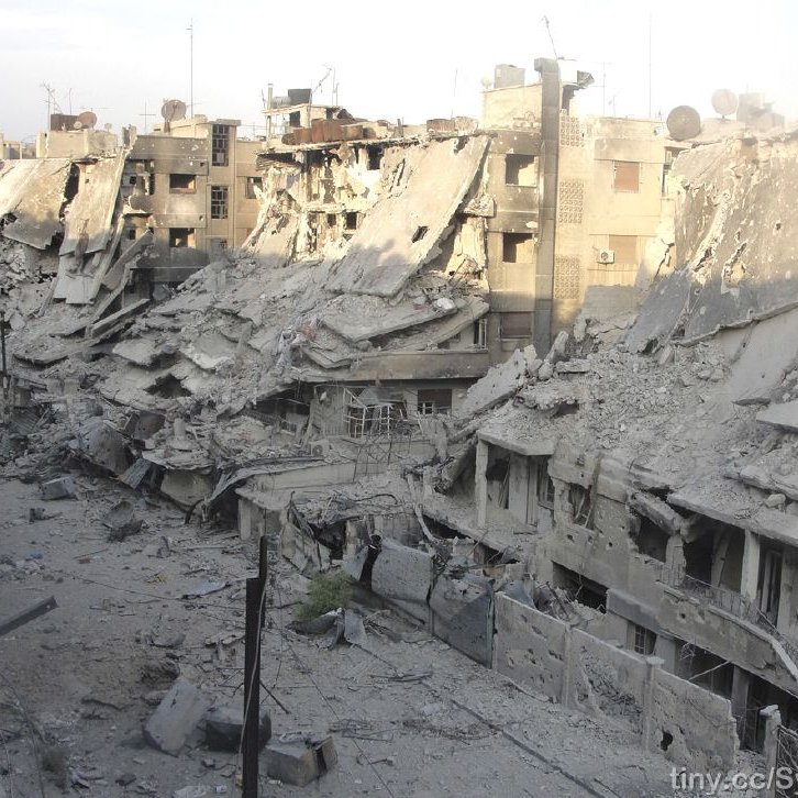 L'image montre un paysage urbain fortement endommagé, avec des bâtiments effondrés et en ruines. Les façades des immeubles sont détruites, laissant entrevoir des débris et des structures instables. Le sol est recouvert de débris et de poussière, témoignant de la violence des destructions. La lumière du jour éclaire la scène, accentuant le contraste entre les ruines et le ciel. Cette image évoque un environnement de guerre et de crise humanitaire.