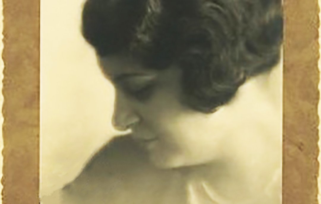 L'image représente un portrait en noir et blanc d'une femme, pris de profil. Elle a les cheveux bouclés et une coiffure élégante typique des années 1920-1930. Son expression est douce et pensive, et elle semble regarder vers le bas, créant une ambiance contemplative. Le fond de l'image est flou, mettant en valeur son visage, et le tout est encadré par une bordure décorative qui ajoute une touche vintage à la photo.