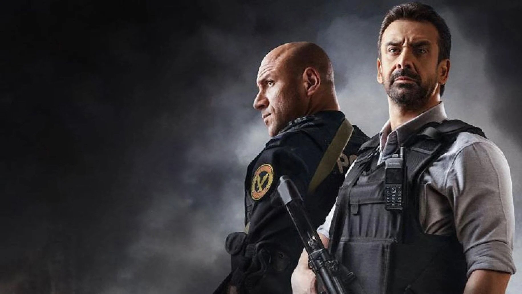 L'image montre deux hommes en tenue tactique, se tenant dos à dos dans une ambiance sombre et intense. L'un d'eux porte un uniforme de police ou militaire, tandis que l'autre est habillé d'une chemise et d'un gilet pare-balles. Ils semblent prêts pour une situation d'urgence, chacun tenant une arme à feu. Le fond est flou et dramatique, ajoutant à la tension de la scène.