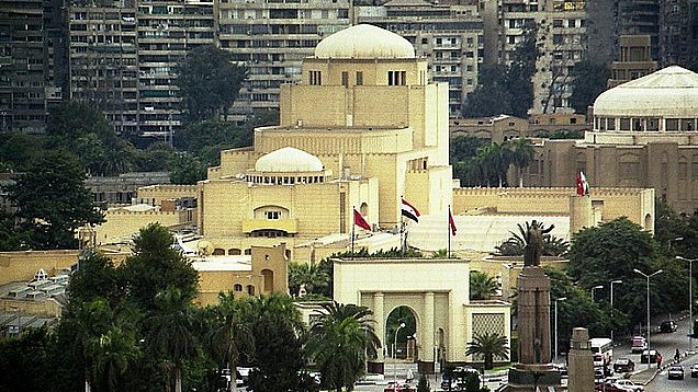 L'image montre un paysage urbain, probablement d'une grande ville. On peut voir des bâtiments aux architectures variées, certains avec des motifs modernes et d'autres avec des influences traditionnelles. Au centre, un bâtiment notable a un style distinctif avec un dôme et des éléments ornementaux, entouré de verdure. Des drapeaux flottent, ce qui suggère l'importance de cet endroit. En arrière-plan, des immeubles d'habitation se dressent, illustrant le contraste entre les différentes époques architecturales.