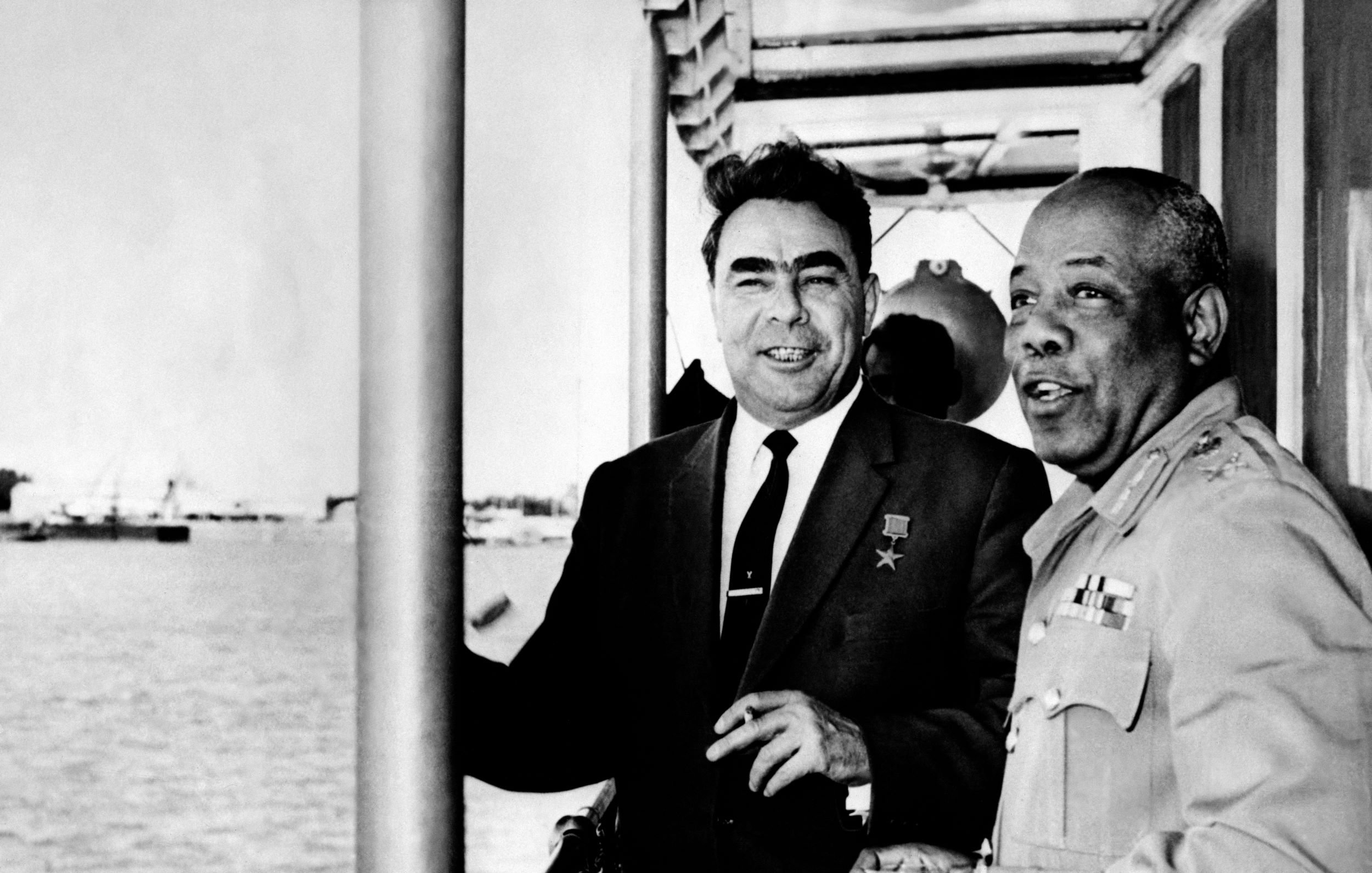 در تصویر، دو مرد در کنار یکدیگر ایستاده‌اند و به نظر می‌رسد که در نزدیکی آب هستند. یکی از آنها لباس رسمی و دکمه‌دار به تن دارد و بر سینه‌اش نشان یا مدالی دیده می‌شود. مرد دیگر نیز لباس نظامی بر تن دارد و در حال صحبت کردن یا تبادل نظر با مرد اول است. پس‌زمینه تصویر نمایی از آب و ممکن است قایق‌ها یا بنادر باشد. احساس کلی تصویر دوستانه و غیررسمی به نظر می‌رسد.