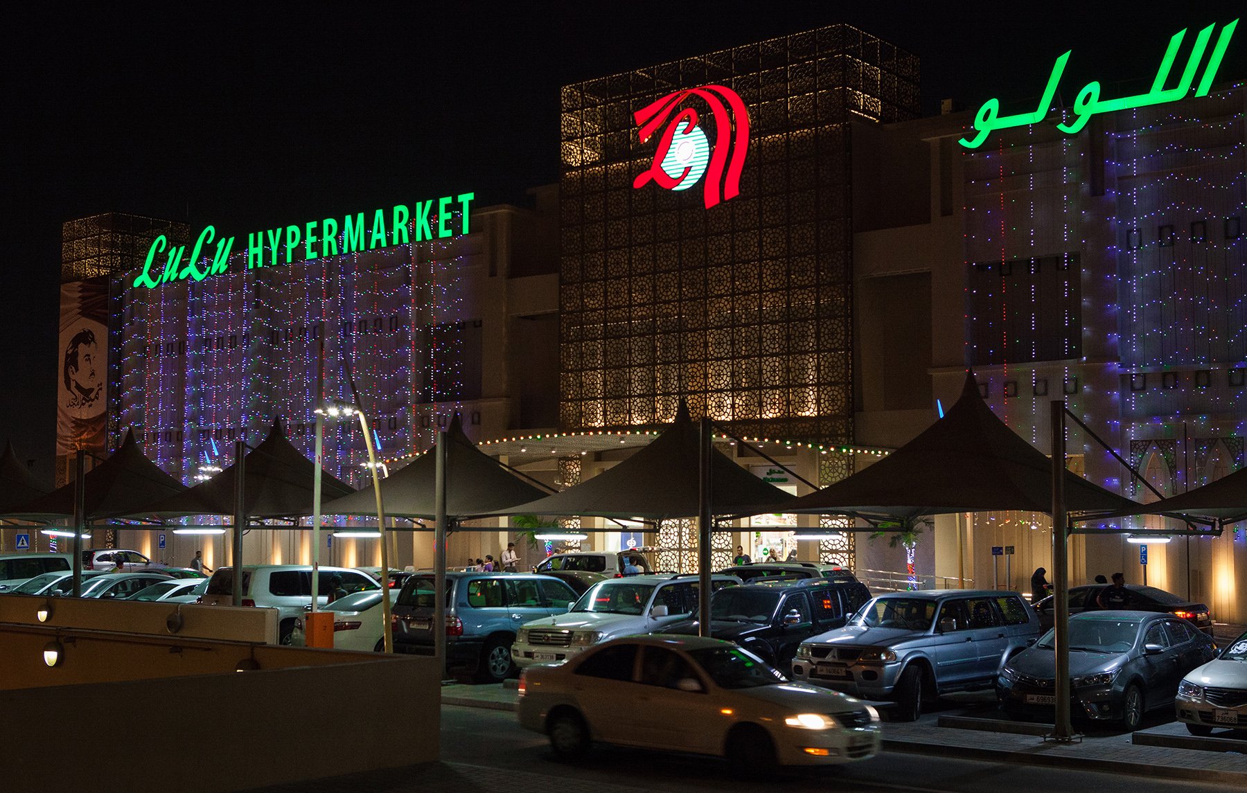 L'image montre un hypermarché, probablement nommé "Lulu Hypermarket", illuminé la nuit. L'extérieur est décoré de lumières, et le nom de l'établissement est affiché en vert, tandis qu'un logo rouge est visible. On aperçoit également un parking rempli de voitures sous des auvents. L'atmosphère paraît animée avec des gens qui semblent entrer ou sortir du magasin.